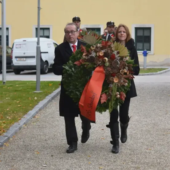 Ein Mann und eine Frau tragen einen Kranz mit rotem Band und Grün. Hinter ihnen geht ein Polizist. Ein weißer Lieferwagen ist geparkt.
