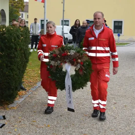 Zwei Personen in roten Uniformen tragen einen Kranz und gehen vor einem Gebäude mit einer Flagge. Dahinter stehen mehrere Personen in der Nähe eines Lieferwagens.