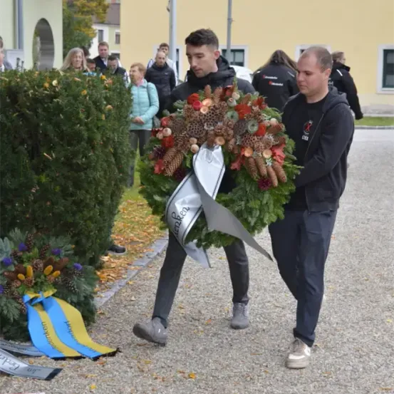 Zwei Männer tragen einen Kranz mit Tannenzapfen und Blumen vor einem Gebäude. Zuschauer stehen herum, einige tragen Jacken. Der Boden ist mit Kies bedeckt.