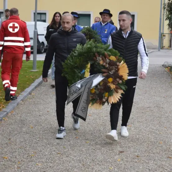 Zwei Männer tragen einen Kranz mit einer Schleife und gehen in einer Prozession. Hinter ihnen geht eine Person in einer roten Uniform, und mehrere andere folgen. Die Umgebung ist im Freien in der Nähe eines Gebäudes.