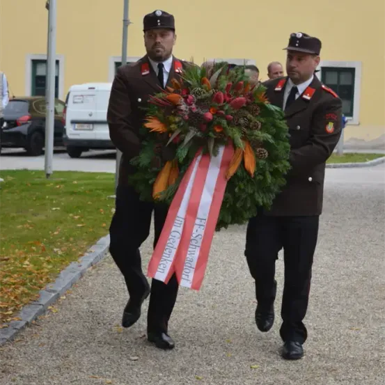 Zwei Feuerwehrleute in Uniform tragen einen Kranz mit einem Band, auf dem 'In Gedenken' steht. Sie gehen auf einem Kiesweg mit einem Gebäude im Hintergrund.