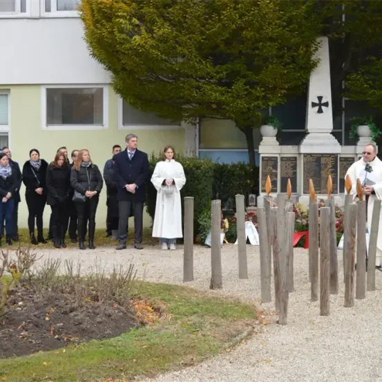 Ein Priester in Weiß steht neben einem Denkmal mit einem Kreuz, umgeben von einer Gruppe von Menschen in Schwarz, einige tragen Schals, in einem Garten mit einem Gebäude und Bäumen im Hintergrund.