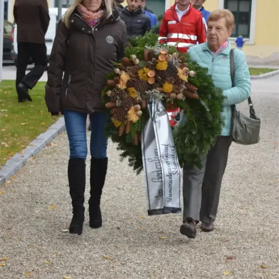 Zwei Frauen tragen einen Kranz mit Tannenzapfen und Blättern auf einem Kiesweg. Eine Frau trägt eine braune Jacke und Stiefel, die andere eine blaue Jacke und Hose. Im Hintergrund gehen Menschen an einem Gebäude vorbei.