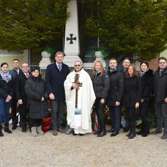 Eine Gruppe von Personen steht vor einer Kirche, wobei ein Priester in der Mitte ein Kreuz hält. Dahinter befindet sich ein Denkmal und ein Gebäude.