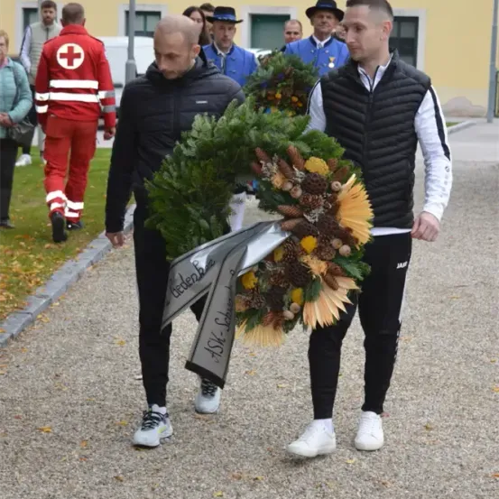 Zwei Männer gehen mit einem Kranz und einer Schleife vorwärts, begleitet von anderen in Uniformen und Jacken. Der Hintergrund zeigt ein Gebäude mit Fenstern und Menschen, die gehen.