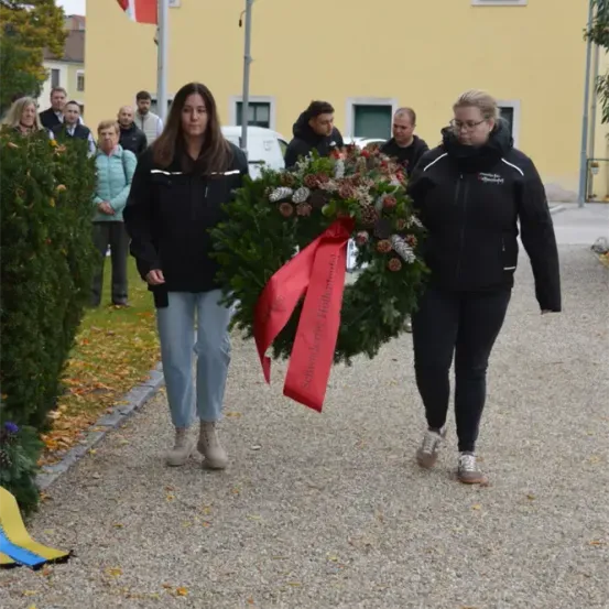 Zwei Frauen in schwarzen Jacken gehen auf einem Kiesweg und tragen einen Kranz. Ein Fahnenmast steht hinter ihnen in der Nähe eines Gebäudes. Mehrere Leute sind in der Nähe versammelt.