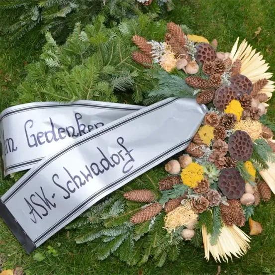 Ein Gedenkkranz mit zwei Bändern auf dem Rasen. Ein Band sagt 'In Gedenken' und das andere 'ASK-Schwadobl'. Der Kranz hat Tannenzapfen, Blätter und Blumen.