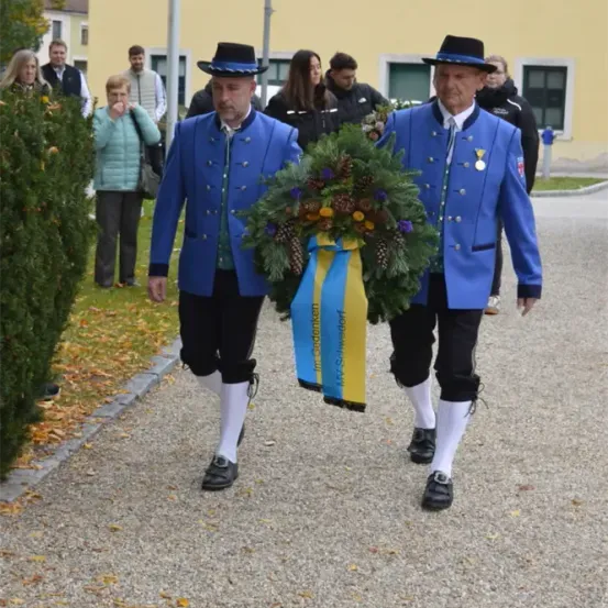 Zwei Männer in blauen Uniformen und Hüten gehen, eine Girlande mit gelben und blauen Bändern tragend. Sie gehen auf einem Kiesweg, mit mehreren Leuten, die hinter ihnen stehen und zusehen.