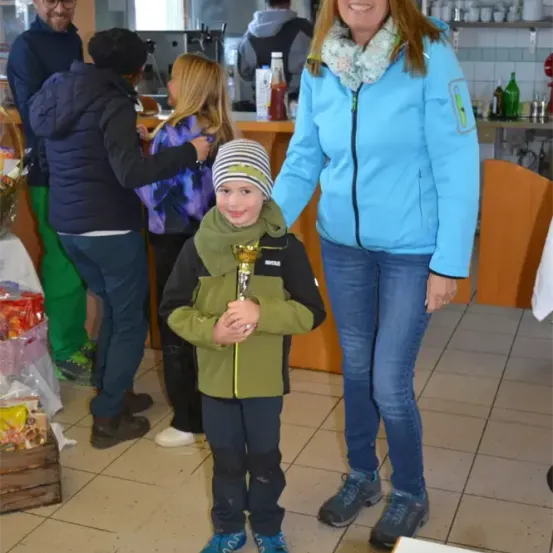 Ein junger Junge mit einem Pokal steht mit einer lächelnden Frau in einem Café mit Menschen im Hintergrund.