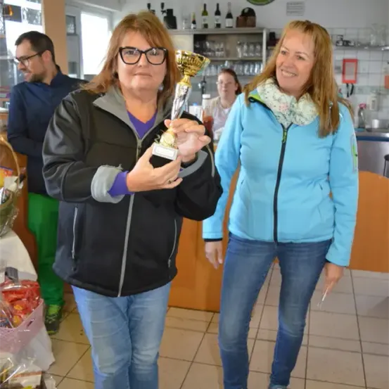 Zwei Frauen stehen in einem Raum. Eine hält eine Trophäe und die andere lächelt und steht neben ihr. Dahinter ist eine Theke mit Regalen. Ein Mann steht hinter der Theke und eine weitere Person sitzt davor.