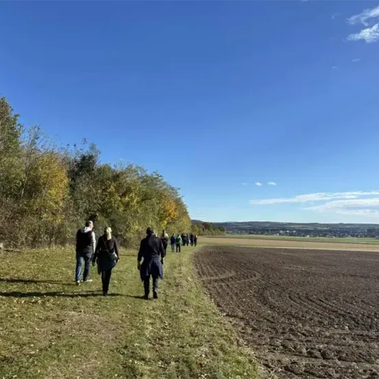 Eine Gruppe von Menschen geht auf einem Grasweg neben einem gepflügten Feld unter einem klaren blauen Himmel.