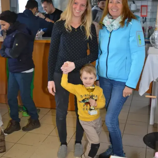 Ein kleiner Junge mit einem Pokal steht lächelnd zwischen zwei Frauen, während eine Gruppe von Menschen hinter ihnen in einem Raum mit gefliesten Böden und einer Theke steht.