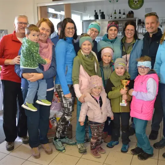 Eine Gruppe von Erwachsenen und Kindern steht lächelnd in einem Raum zusammen. Eine Frau hält ein Kind, und ein Mann hält eine Trophäe. Sie tragen alle Winterkleidung.