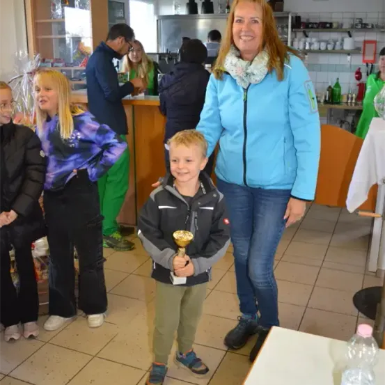 Eine Frau und ein Junge, die eine Trophäe halten, stehen zusammen. Der Junge trägt eine Jacke und Turnschuhe. Dahinter schaut ein Mädchen zur Kamera. Es gibt einen Tisch mit einer Flasche und ein Regal mit Gegenständen.