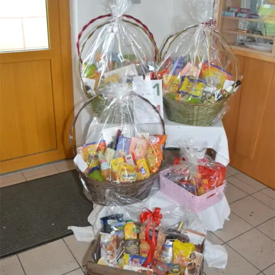Drei Körbe mit verschiedenen Snacks stehen auf einem Tisch, jeder in Plastik verpackt. Die Körbe haben unterschiedliche Größen und Farben, einer hat ein rotes Band. Im Hintergrund sind eine Holztür und ein Regal mit Gegenständen zu sehen.