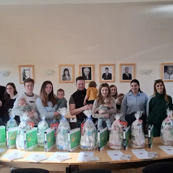 Eine Gruppe von Menschen posiert für ein Foto mit verpackten Geschenken und Babys vor einer Wand mit gerahmten Porträts.