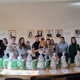 Eine Gruppe von Menschen posiert für ein Foto mit verpackten Geschenken und Babys vor einer Wand mit gerahmten Porträts.