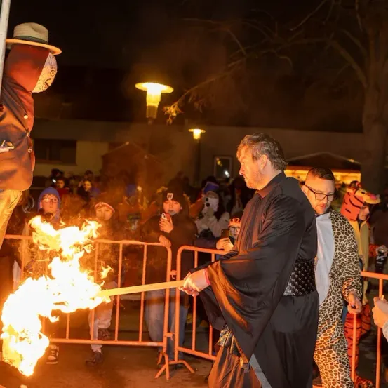 Eine Gruppe von Menschen versammelt sich bei Nacht draußen und beobachtet einen Mann in einem Gewand, der eine Fackel mit Feuer schwingt. Er scheint eine Zeremonie oder einen Ritus durchzuführen.