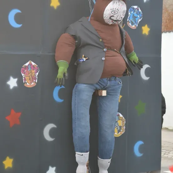 Eine Puppe mit bemaltem Gesicht und Hut, gekleidet in einer Jacke, Jeans und Stiefeln, hängt an einer Kette an einem schwarzen Hintergrund mit verschiedenen Symbolen.