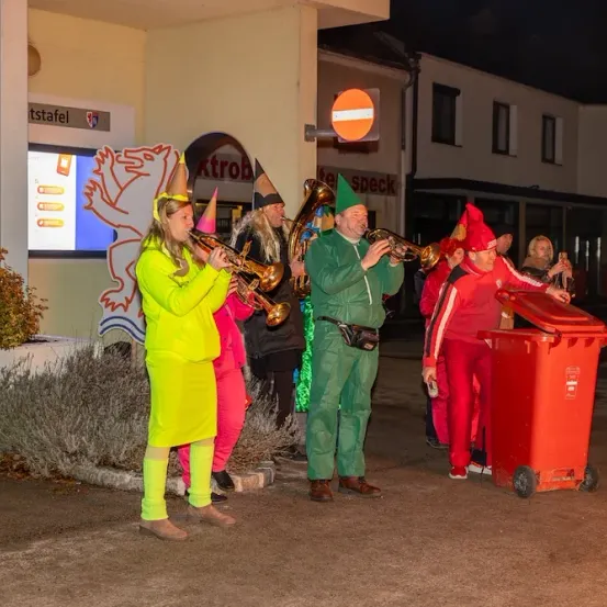 Eine Gruppe von Menschen in bunten Kostümen spielt Blasinstrumente vor einem Gebäude. Der Mann in Grün bläst eine Trompete, und eine Frau in Gelb spielt eine Posaune.