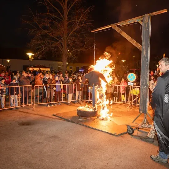 Ein Mann in einer Robe steht neben einem brennenden Feuerkorb, umgeben von einer Menge bei Nacht.