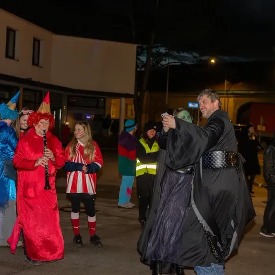 Eine Gruppe von Menschen in Kostümen, die nachts Musikinstrumente spielen. Ein Mann in einem schwarzen Umhang lächelt und macht ein Selfie.