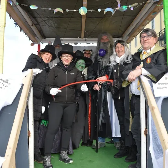 Eine Gruppe von Menschen in Kostümen steht unter einem Vordach auf einer Straße. Eine Frau hält eine rote Schnur. Ein Schild zeigt 'An Jugendliche jung kein ausges.' Ein Mann im Gewand hält einen Stock.