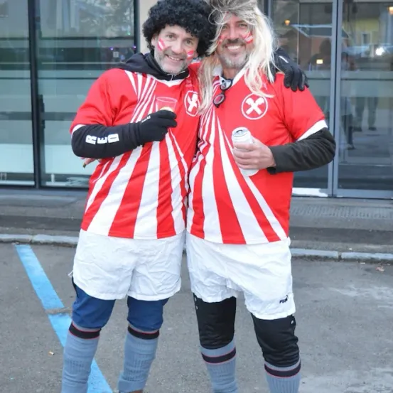 Zwei Männer in roten und weißen gestreiften Trikots stehen nebeneinander auf einem Parkplatz. Einer hält eine Dose. Sie haben Farbe im Gesicht. Hinter ihnen ist ein Glasgebäude.