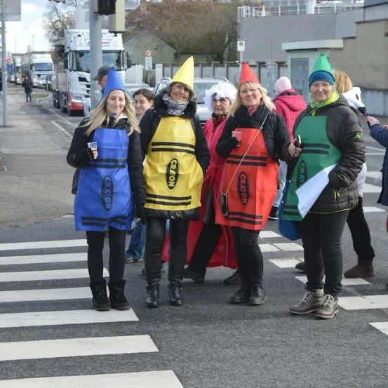 Fünf Frauen in Schürzen und Hüten, verkleidet als Buntstifte, stehen auf einem Zebrastreifen mit einem Fußgängerübergangsschild. Ein Mann macht ein Foto.
