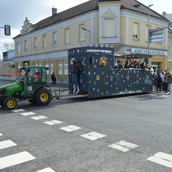 Ein geschmückter Traktor zieht einen Festwagen in einer Parade. Leute reiten auf dem Wagen, und andere gehen am Straßenrand. Im Hintergrund sind Gebäude und Straßenlaternen zu sehen.