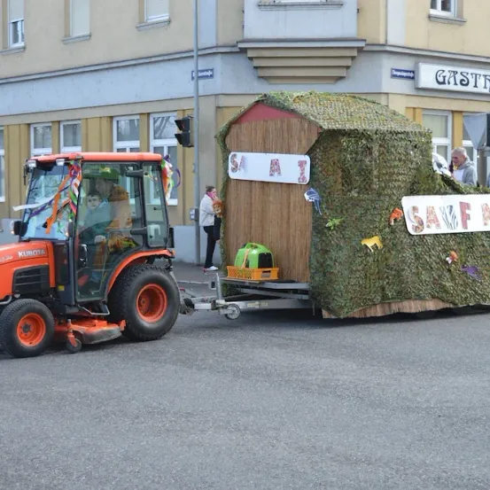 Ein Traktor zieht einen geschmückten Anhänger mit einem hausartigen Aufbau, der mit Moos und Tierfiguren bedeckt ist. Menschen gehen hinter dem Anhänger her.