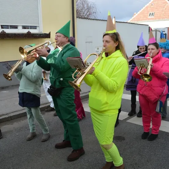 Eine Gruppe von Musikern in Kostümen spielt Trompeten auf der Straße. Sie tragen leuchtende Outfits und Kegelhüte. Hinter ihnen gehen Menschen auf dem Bürgersteig.