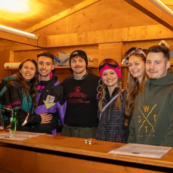 Eine Gruppe von sechs Menschen in Winterkleidung posiert lächelnd für ein Foto in einem hölzernen Raum. Dahinter sind Regale mit Jacken und Helmen gefüllt. Vor ihnen befindet sich eine Bartheke.