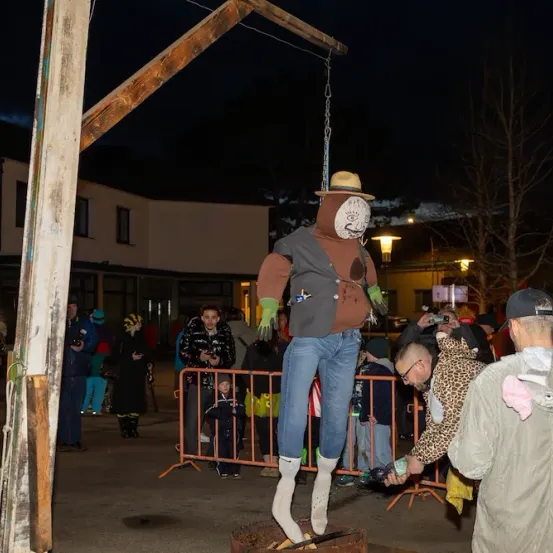 Eine Menschenmenge hat sich um eine Vogelscheuche versammelt, die von einem Baum hängt. Die Vogelscheuche trägt einen Hut, Handschuhe und Stiefel. Die Menge beobachtet und einige machen Fotos. Im Hintergrund befinden sich Gebäude und Bäume.