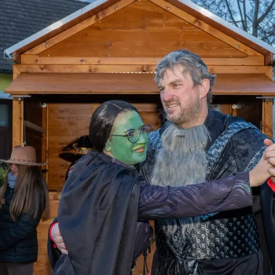 Ein Mann und eine Frau in Kostümen, möglicherweise für Halloween, lächeln und umarmen sich vor einer Holzstruktur. Die Frau trägt einen schwarzen Umhang und grünes Gesichtsmake-up. Hinter ihnen ist eine weitere Frau mit Hut und langen Haaren zu sehen.