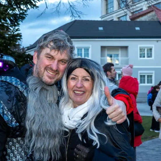 Ein Mann und eine Frau in Kostümen lächeln in die Kamera. Dahinter gehen Menschen spazieren, und ein Gebäude mit rotem Dach ist sichtbar.