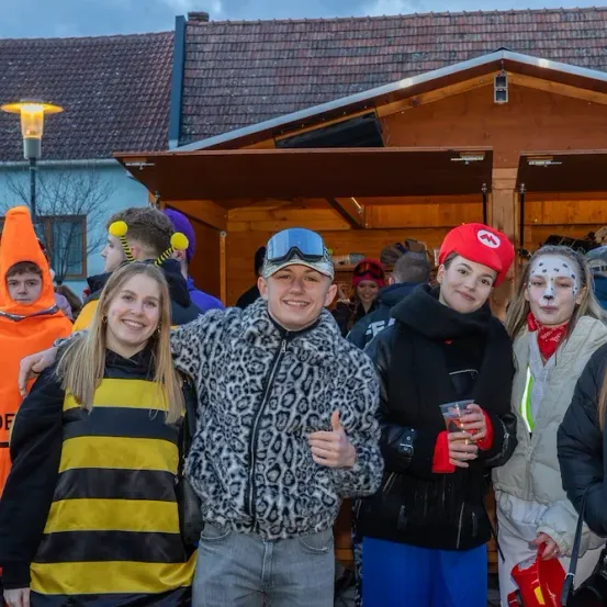 Eine Gruppe von Freunden in Kostümen steht vor einer Holzbude. Einige haben Gesichtsbemalung, und einer hält einen Becher. Das Wetter ist kühl.