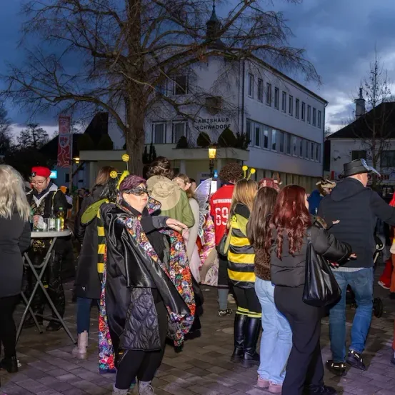 Eine Gruppe von Menschen in verschiedenen Kostümen versammelt sich auf einem Stadtplatz, eine Frau zeigt auf etwas vor ihnen. Ein Gebäude und ein Baum sind im Hintergrund zu sehen.