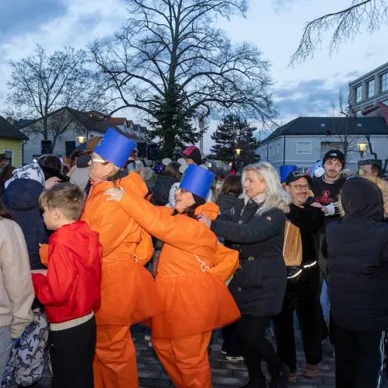 Eine Gruppe von Menschen in orangefarbenen Anzügen und blauen Hüten versammelt sich auf einer Straße für eine Veranstaltung, umgeben von Gebäuden und Bäumen unter einem bewölkten Himmel.
