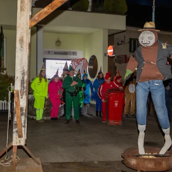 Eine Gruppe von Menschen in verschiedenen Kostümen steht vor einem Gebäude. Ein Mann in einem Heuballen-Kostüm steht auf einer runden Plattform.