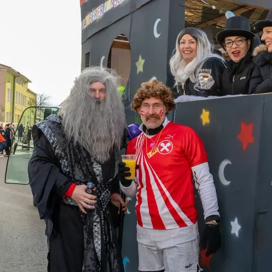 Zwei Männer in Kostümen posieren vor einer dekorierten Wand. Einer in einem Gandalf-Kostüm hält ein Getränk, während der andere in einem rot-weißen Trikot ein Bier hält. Eine Gruppe von Menschen steht hinter ihnen und hält Getränke. Im Hintergrund sind Gebäude und Bäume zu sehen.
