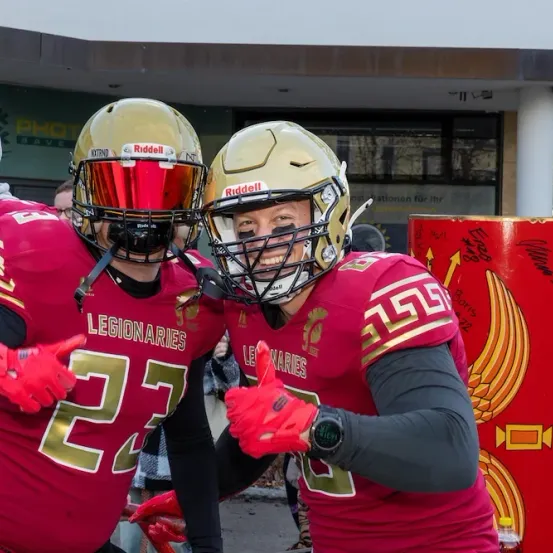 Zwei Footballspieler in roten Uniformen mit goldenen Helmen posieren für ein Foto vor einem Gebäude. Einer trägt die Nummer 27 und der andere die Nummer 25. Sie lächeln und zeigen Daumen hoch. Ein rotes Objekt mit goldenen Designs steht hinter ihnen.