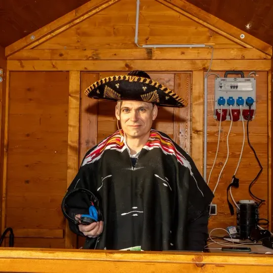 Ein Mann in einem Sombrero steht in einem Holzraum und hält ein Gerät in den Händen. Hinter ihm befindet sich eine Holztür und eine Steckdose.