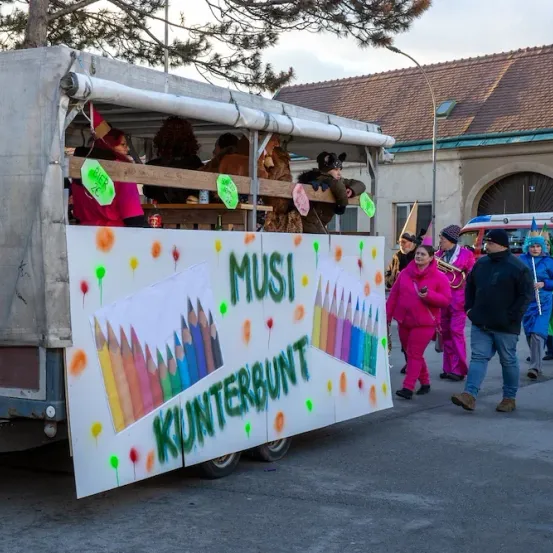Ein bunter Umzugswagen mit Menschen im Inneren und einem Schild mit bunten Stiften und dem Wort Mus.