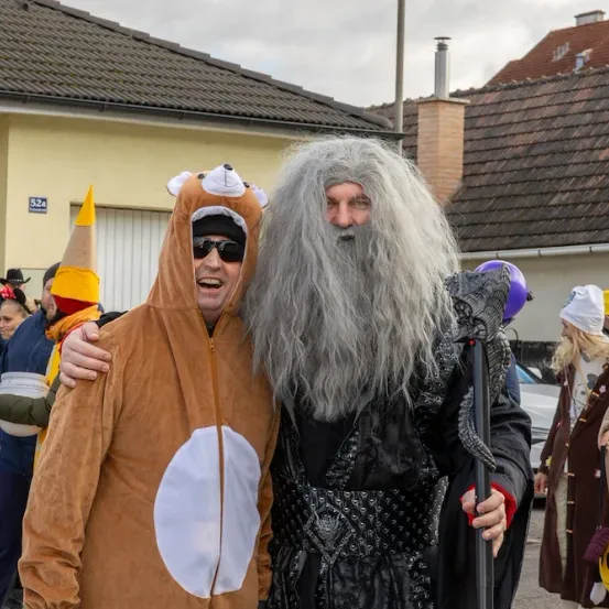 Zwei Menschen in Kostümen stehen draußen, einer in einem Bärenkostüm und der andere in einem Gandalf-ähnlichen Kostüm, mit Gebäuden und geparkten Autos im Hintergrund.