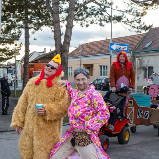 Drei Männer in Kostümen posieren auf der Straße. Einer fährt einen Spielzeugtraktor mit einem Schwein in einem hausförmigen Anhänger. Ein blaues Straßenschild weist auf Einbahn hin.