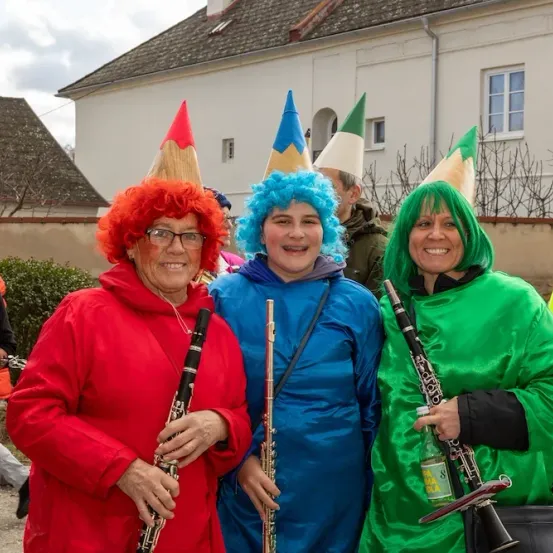 Drei Frauen in farbenfrohen Kostümen posieren mit Musikinstrumenten vor einem weißen Gebäude mit Schornstein und Kamin.