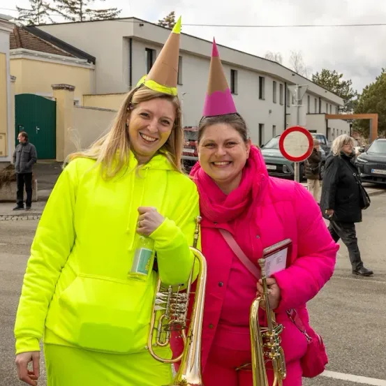 Zwei Frauen in leuchtenden Neon- und Pink-Outfits stehen draußen, lächeln und halten Trompeten. Eine trägt einen Partyhut. Im Hintergrund sind Gebäude, geparkte Autos und Menschen zu sehen.
