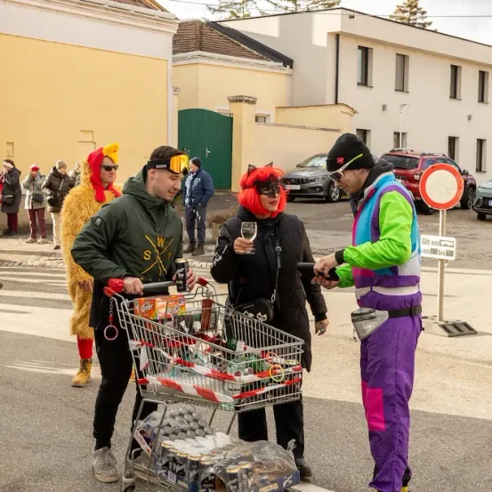 Eine Straßenszene mit Menschen in Kostümen, ein Mann mit einem gefüllten Einkaufswagen und ein weiterer Mann mit einem Weinglas. Im Hintergrund sind Gebäude, geparkte Autos und Menschen zu sehen, die gehen.