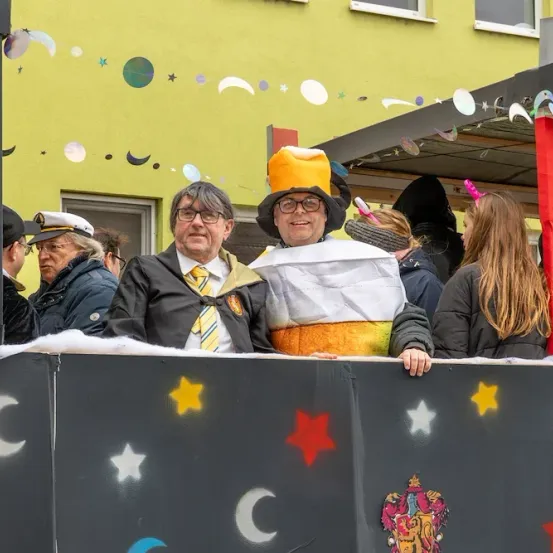 Zwei Männer in Kostümen, einer als Zauberer verkleidet, stehen auf einem geschmückten Wagen mit bemalten Sternen und Monden. Dahinter beobachten Menschen in verschiedenen Kostümen von einem Gebäude mit gelben Wänden aus.